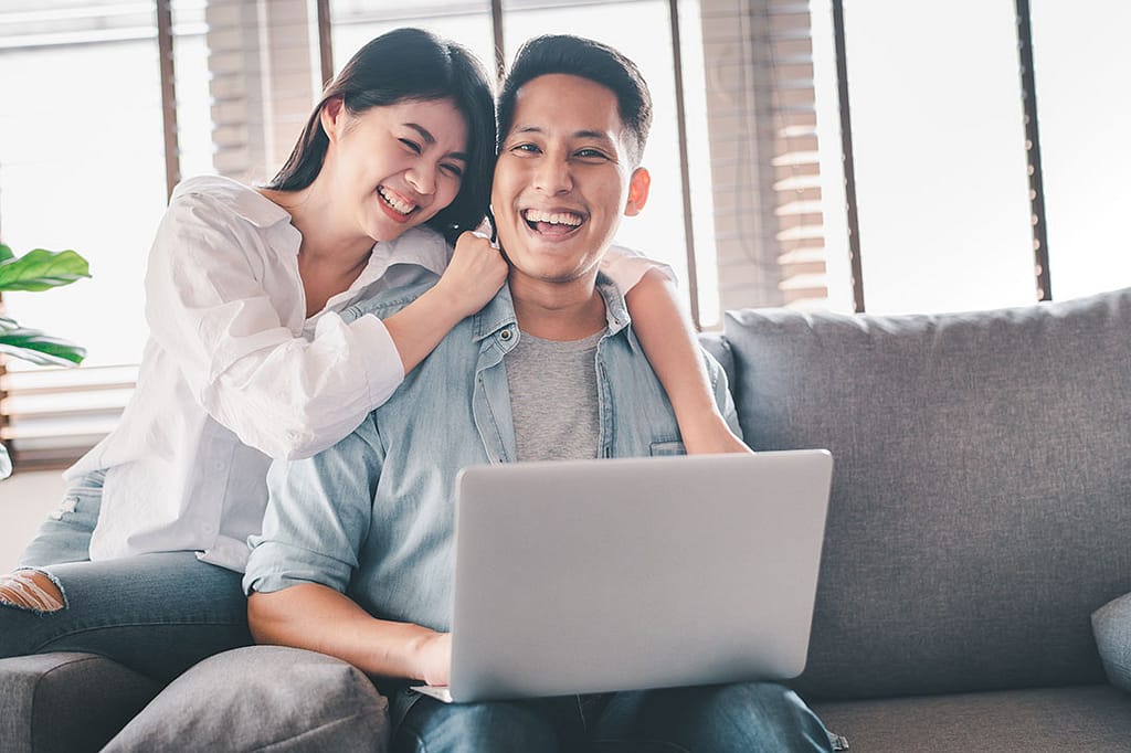 happy smiling asian couple on the couch looking at a laptop