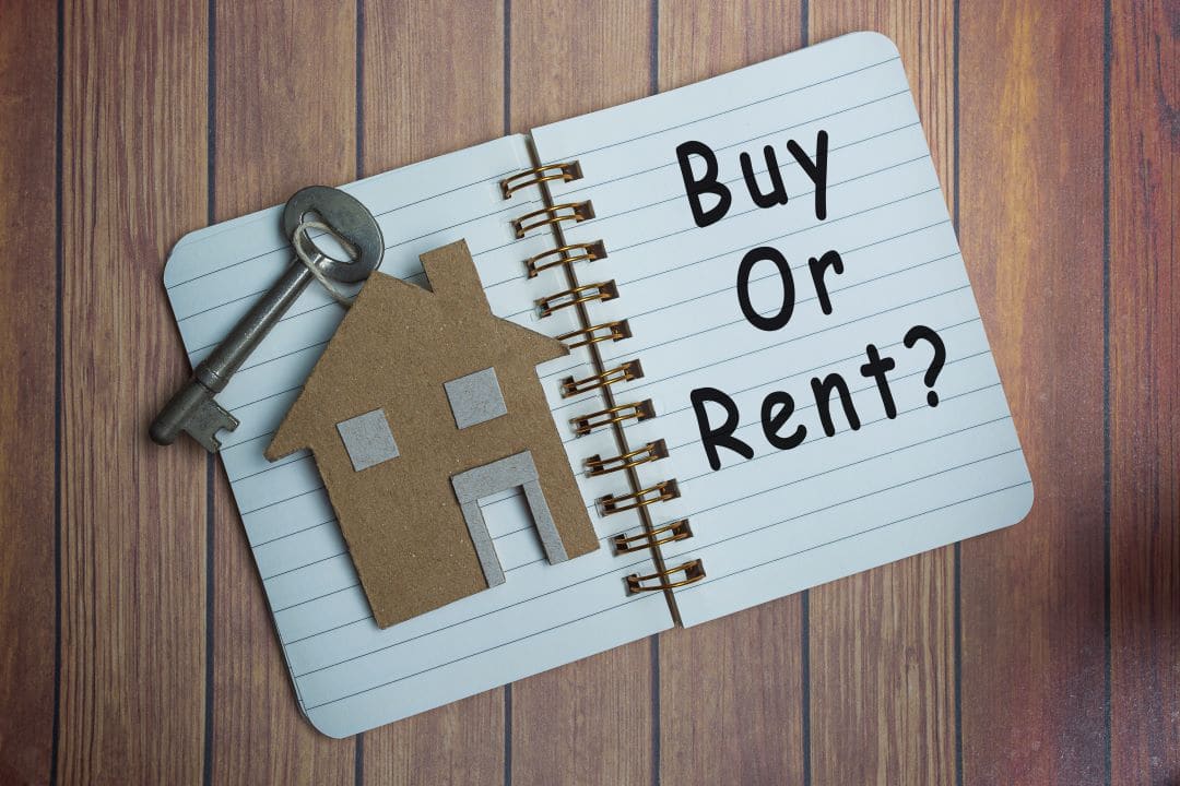 A cardboard cutout of a house with a metallic old looking key sit on a notepad with it open. On the other page are the words, "Buy or rent?".